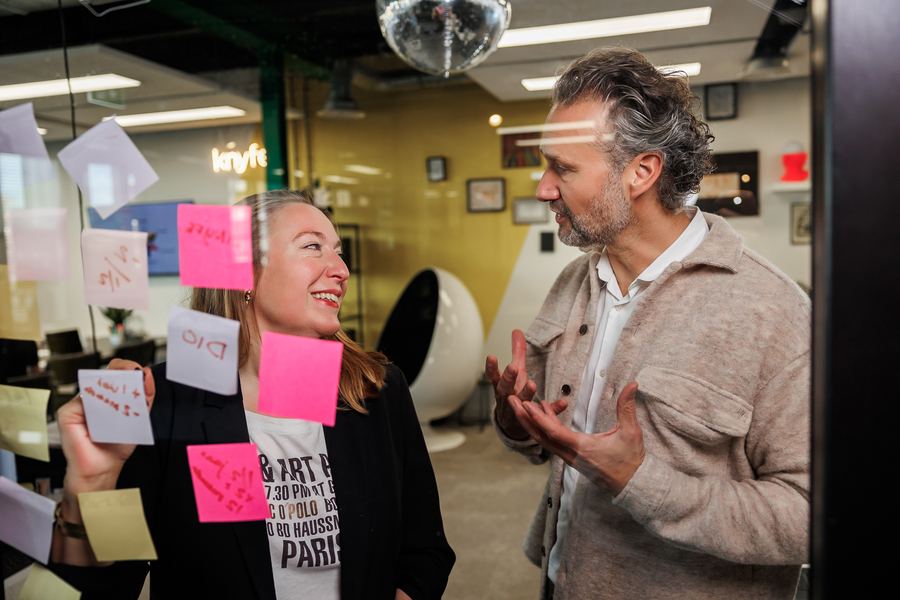 Two colleagues mid-discussion behind a wall of sticky notes, a mirror ball above