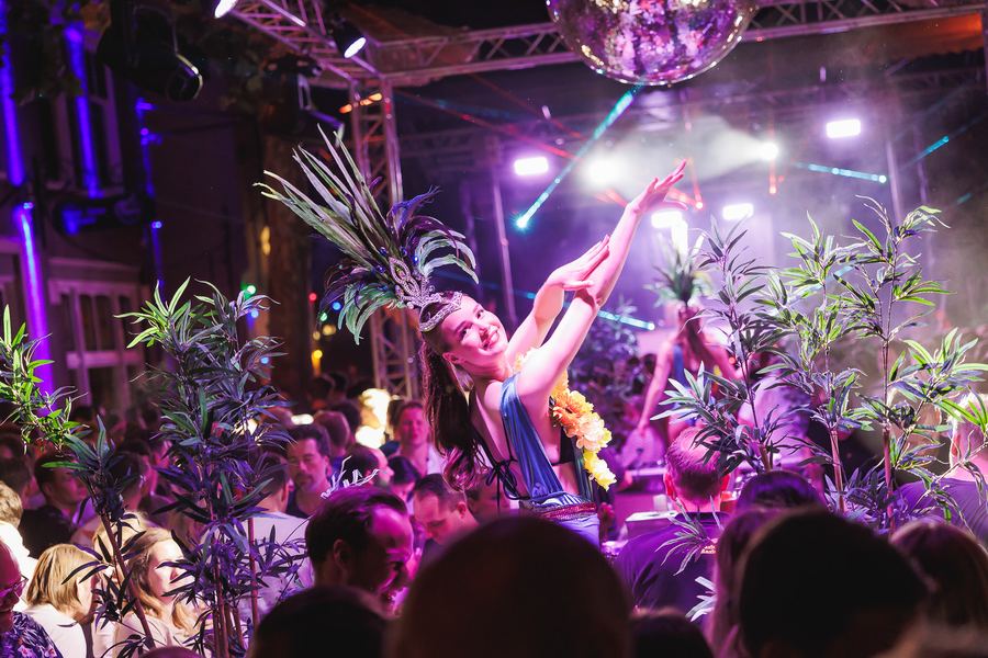 A dancer on a podium among palm leaves, arms stretched to the lights