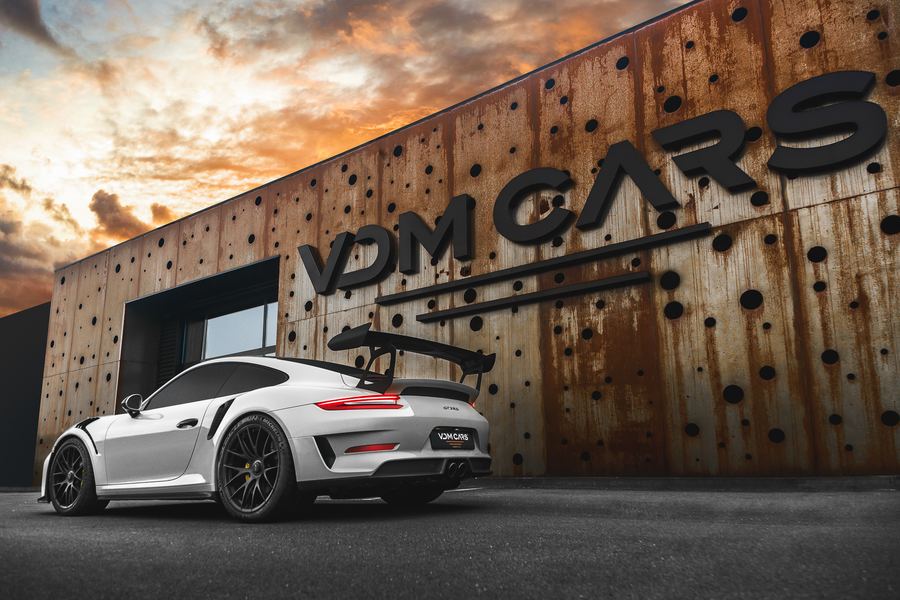 A white Porsche 911 with a rear wing, parked at VDM Cars’ rusted steel facade