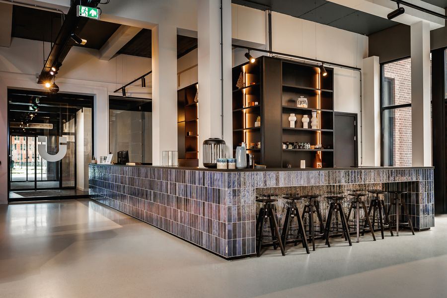A bar faced in blue-grey tiles, a row of stools, open shelving behind
